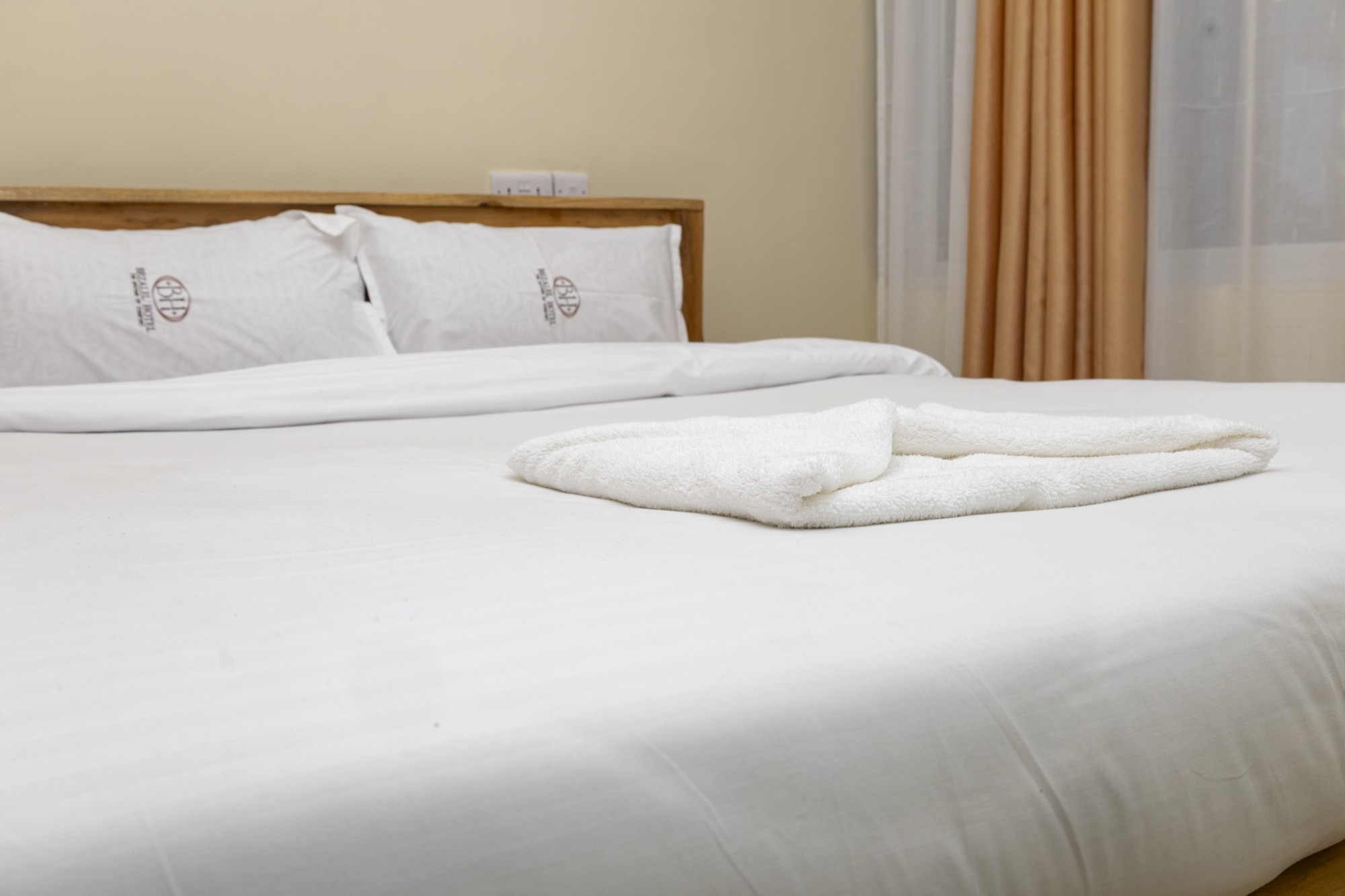 A made-up 5 by 6 bed with a folded towel, curtains drawn back